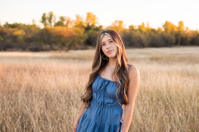 senior girl with long brown hair wearing a blue sundress stands in tall grass with trees behind her smiling softly in a country field in the fall near Oklahoma City