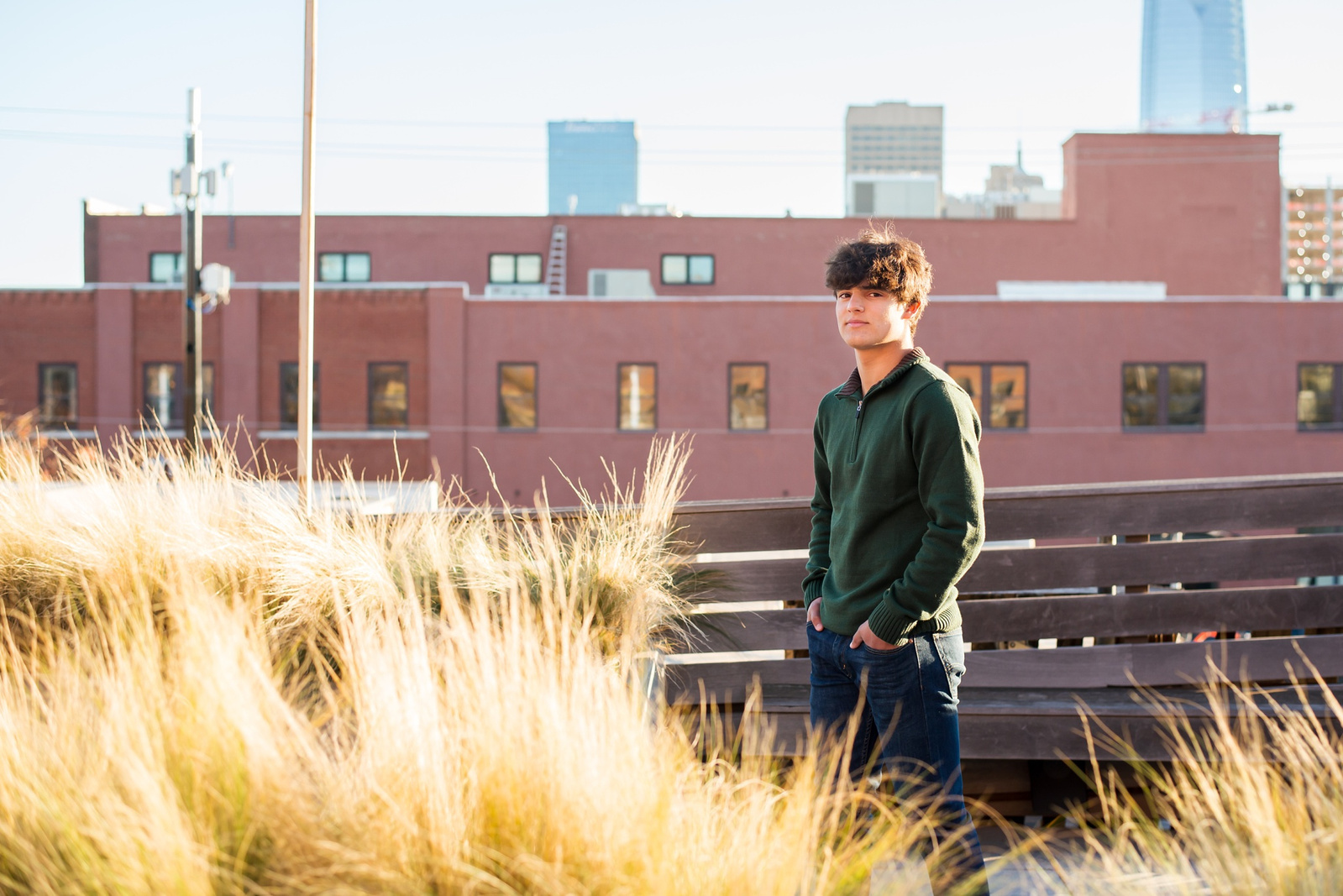 high school senior boy with dark hair wearing green sweater and jeans stands on a rooftop with gold grass around him and the OKC downtown skyline behind him