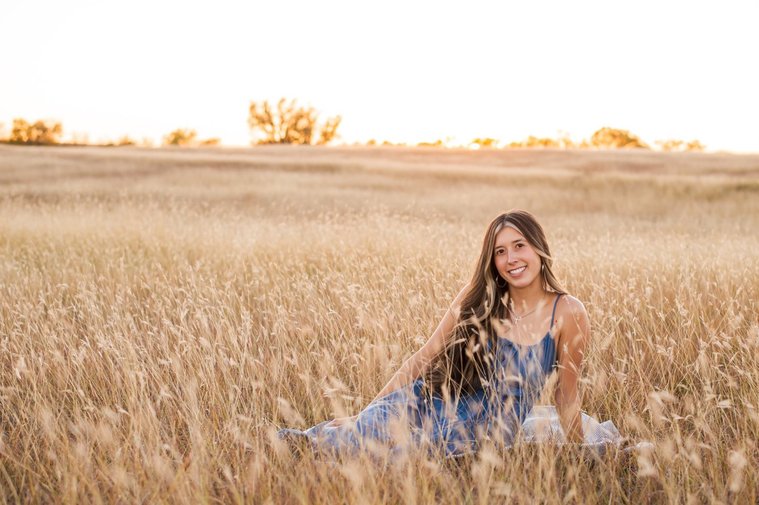 senior girl with long brown hair wearing a blue sundress sits among tall golden grass smiling in a country field in the fall near Oklahoma City