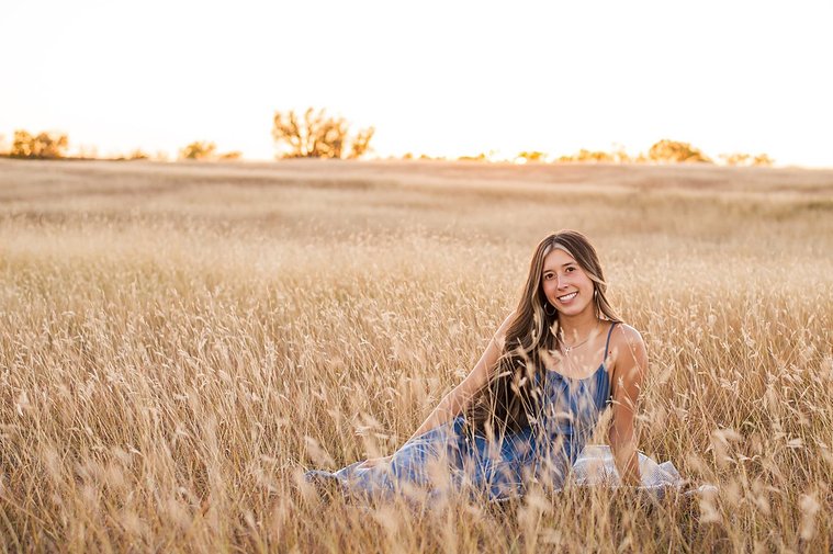 senior girl with long brown hair wearing a blue sundress sits among tall golden grass smiling in a country field in the fall near Oklahoma City