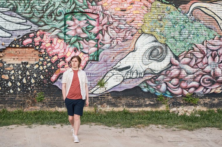 senior boy wearing a button down shirt and blue shorts walks toward camera in front of a wall mural in midtown Oklahoma City
