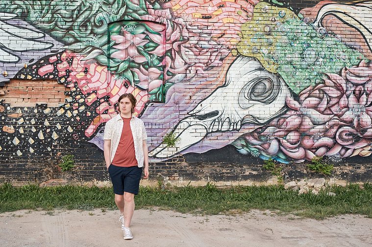 senior boy wearing a button down shirt and blue shorts walks toward camera in front of a wall mural in midtown Oklahoma City