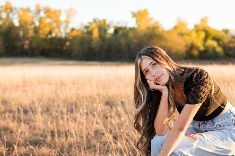 senior girl with long brown hair wearing a black top and jeans sits on a stool in tall grass with trees behind her, with her chin in hand looking softly at camera in a country field in the fall near Oklahoma City