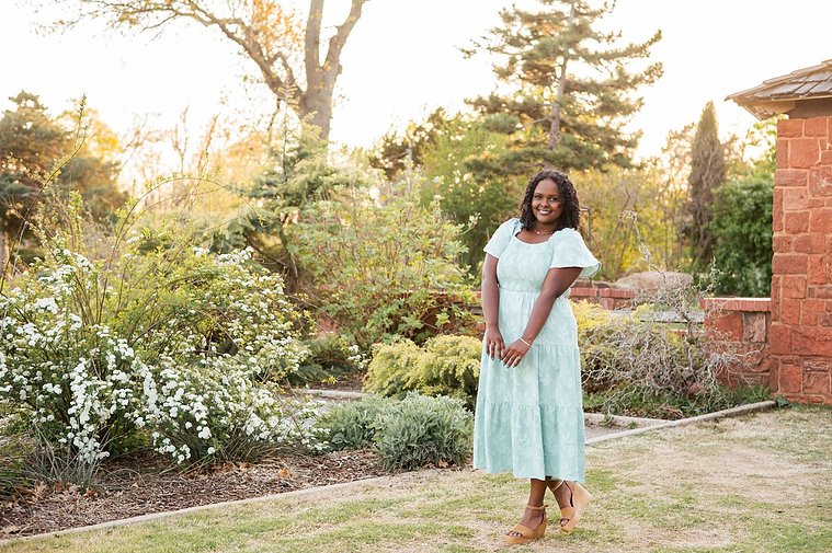 senior girl wearing a dress stands prettily and smiles among the flowers and trees of Will Rogers Gardens in OKC