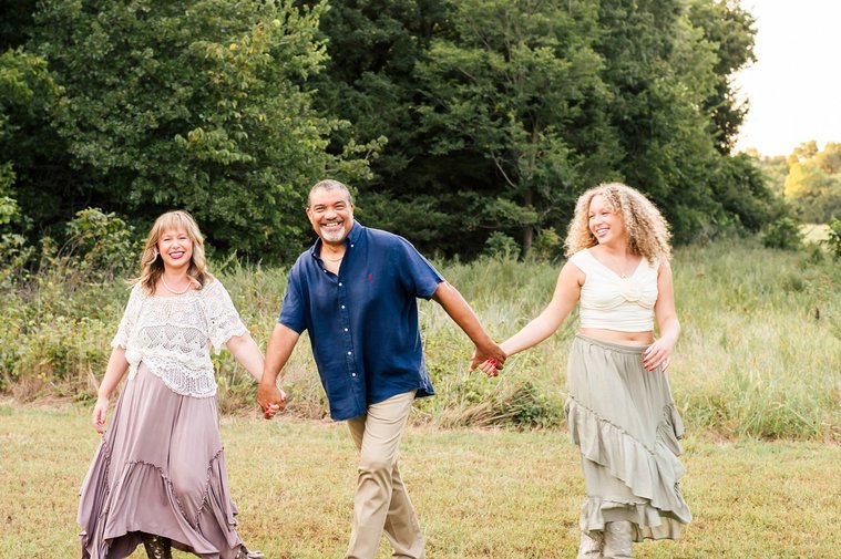 a mom, dad, and teen daughter hold hands in a line walking and laughing togther in a field with trees behind them in Norman, Oklahoma