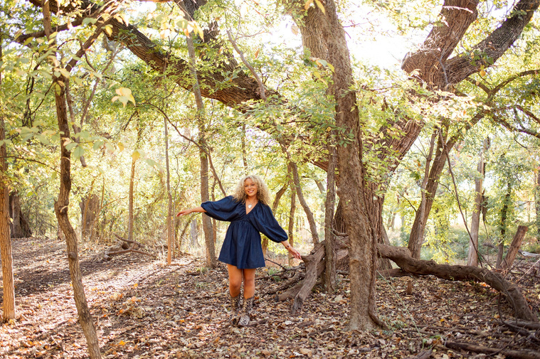 wide angle portrait of a high school senior girl with blonde curly hair and blue eyes wearing a blue dress and cowboy boots in front of a large tree at Martin Nature Park in Oklahoma City
