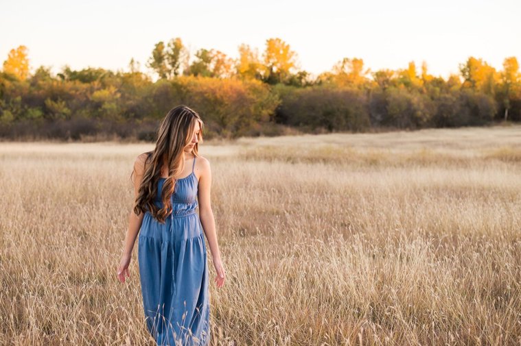 senior girl with long brown hair wearing a blue sundress walks in a country field in the fall with tress behind her and looking down softly near Oklahoma City