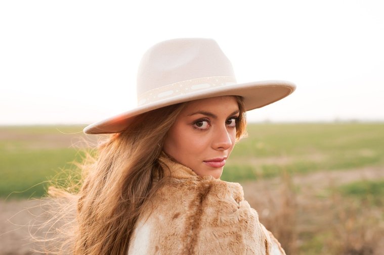 high school senior girl with long brown hair and beautiful eyes, wearing a western hat wrapped in a brown blanket looks over her shoulder with a serious expression standing in a field near OKC with the sunset behind her