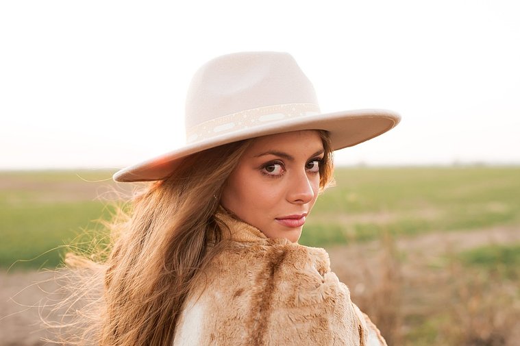 high school senior girl with long brown hair and beautiful eyes, wearing a western hat wrapped in a brown blanket looks over her shoulder with a serious expression standing in a field near OKC with the sunset behind her