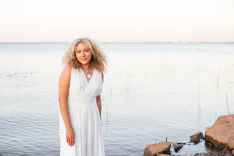 portrait of a high school senior girl in a white flowy dress standing in the water swishing her skirt and smiling on the shore of Lake Hefner in Oklahoma City