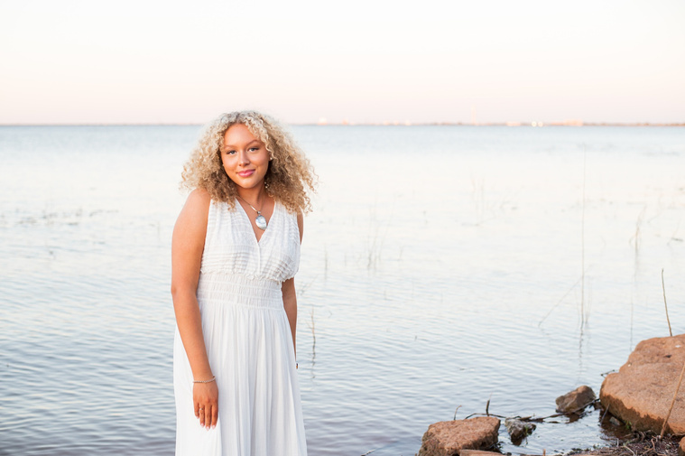 portrait of a high school senior girl in a white flowy dress standing in the water swishing her skirt and smiling on the shore of Lake Hefner in Oklahoma City