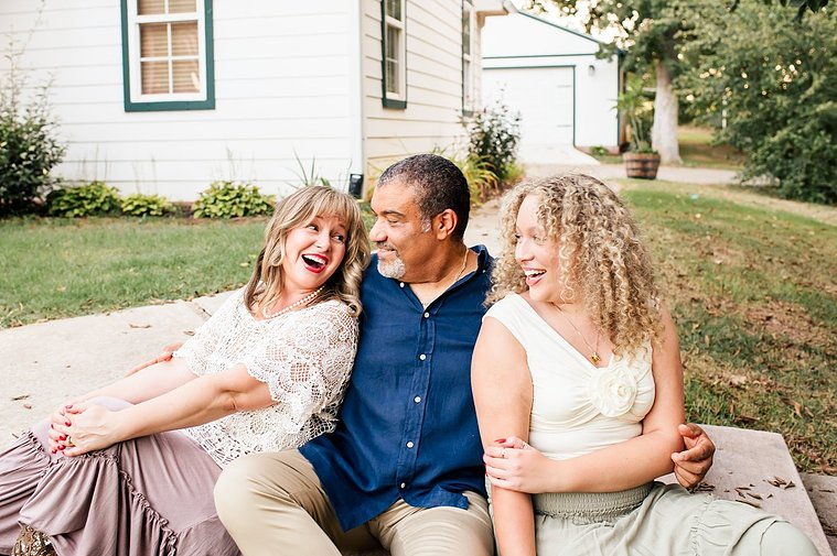 a mom, dad, and teen daughter sit together hugging and laughing on their front sidewalk with their house behind them in Norman, Oklahoma