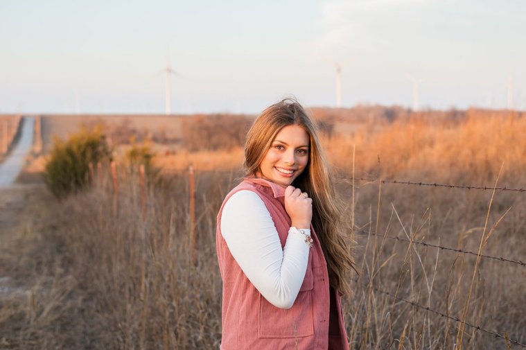 beautiful senior girl with long hair wearing a pink vest smiling with one hand on her collar with a country road and fields behind her in Oklahoma