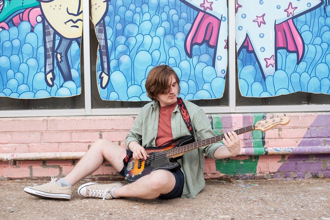 senior boy in shorts and a button down sits against a wall mural in Oklahoma City holding a bass guitar and looking at it