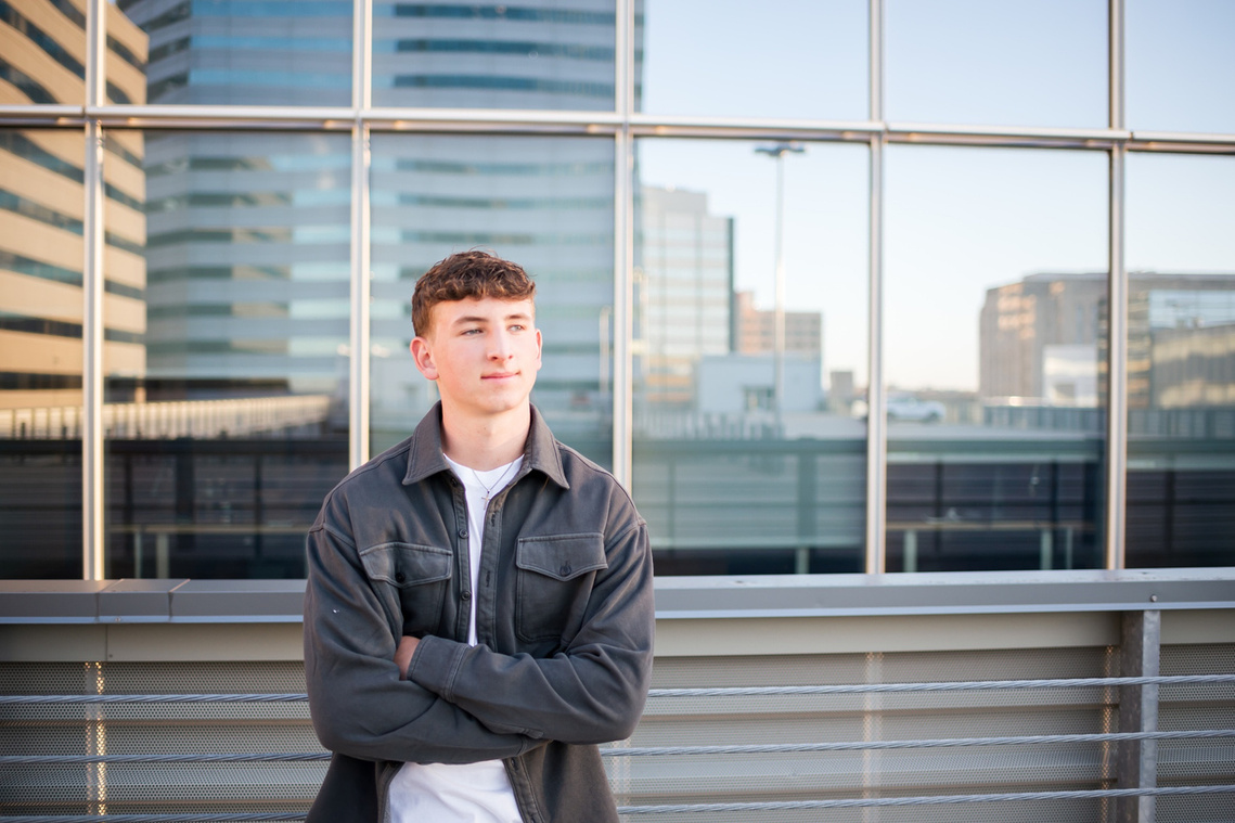 high school senior boy wearing a gray jacket and white t-shirt crosses his arms and looks off to the side standing on a parking garage rooftop with downtown OKC buildings reflected behind him