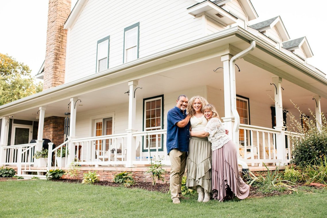 a mom, dad, and teen daughter stand together hugging and smiling in front of their two story home with a wraparound porch in Norman, Oklahoma