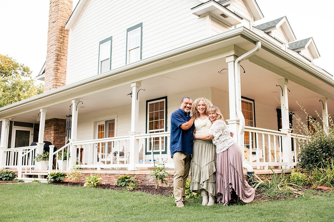 a mom, dad, and teen daughter stand together hugging and smiling in front of their two story home with a wraparound porch in Norman, Oklahoma