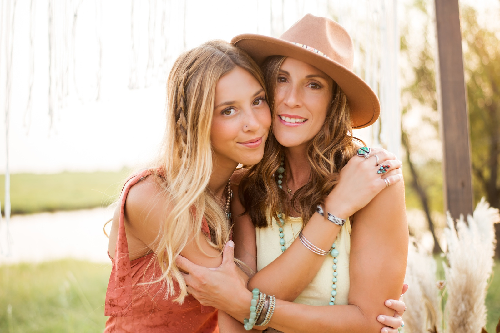 a teen daughter and her mom hug each other wearing boho clothing and accessories with pampas grass and macrame around them in a field with a poind in central Oklahoma