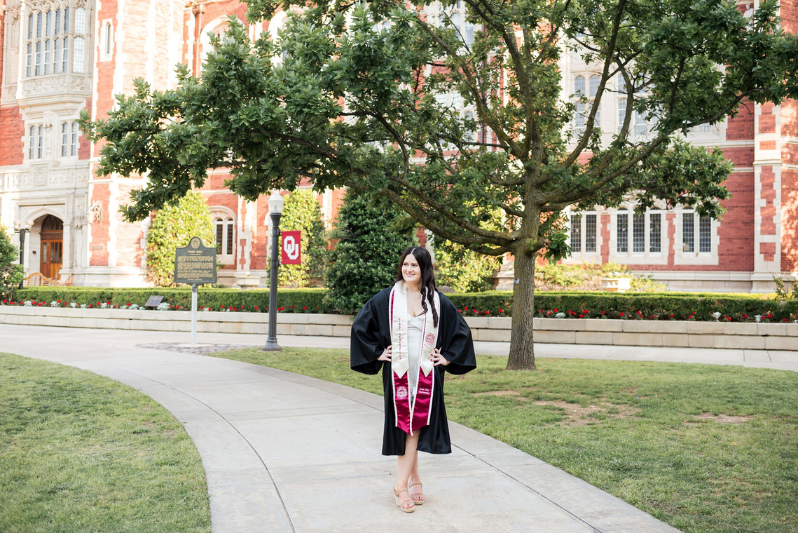 college senior girl wearing black gown and stole stands on a sidewalk in the Parrington Oval on the OU campus in Norman with Evans Hall behind her