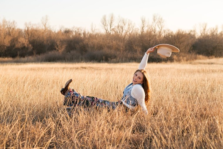 high school senior girl with long brown hair wearing cowboy boots, Wranglers, a denim vest and western hat smiles over her shoulder and holds her hat in the air as she sits in a field of golden grass in the country near OKC