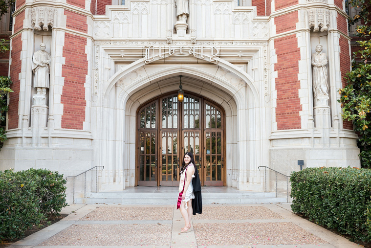 wide angle portrait of a  college senior girl wearing white dress and stole stands holding her black gown over one shoulder on a sidewalk in front of Bizzell Library on the OU campus in Norman with the main door behind her