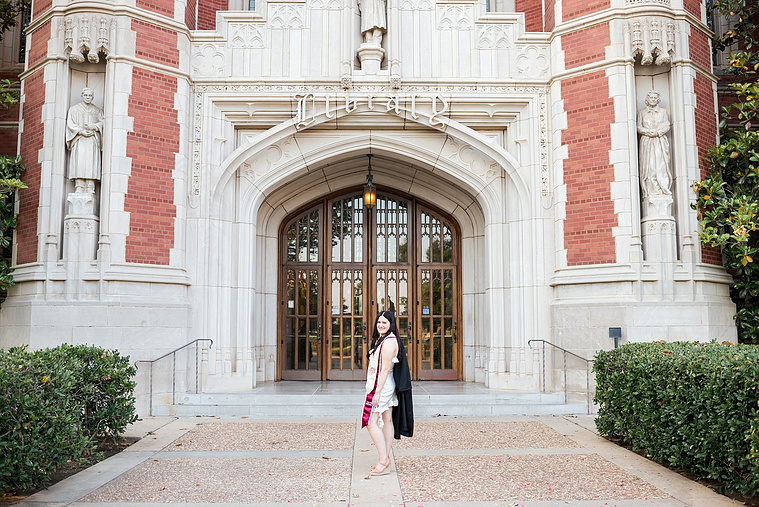 wide angle portrait of a  college senior girl wearing white dress and stole stands holding her black gown over one shoulder on a sidewalk in front of Bizzell Library on the OU campus in Norman with the main door behind her