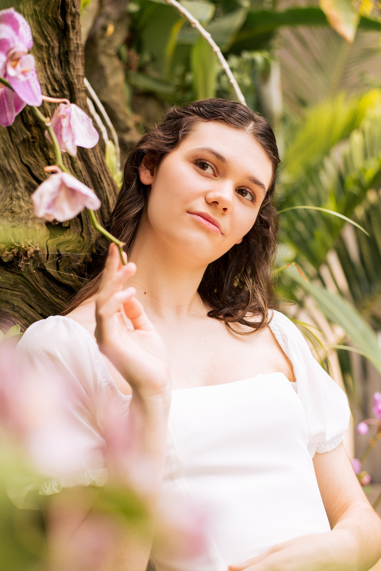 high school senior girl with long brown hair in a white blouse holds the end of a purple flower against a tropical plant wall while she looks up dreamily inside the Crystal Bridge at Myriad Gardens in Oklahoma City 