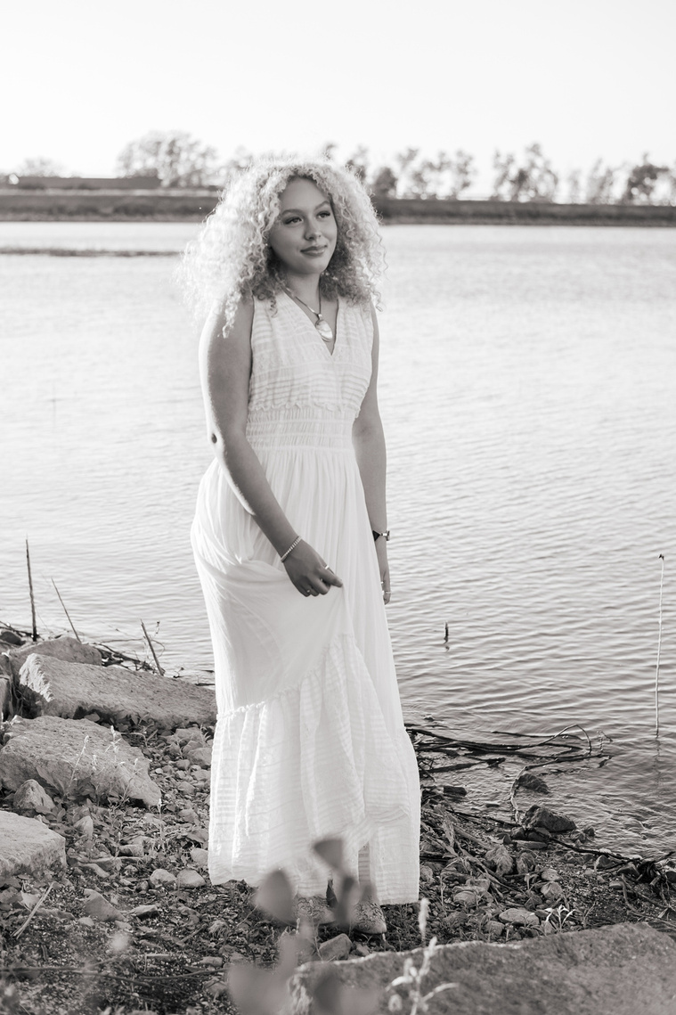 black and white vertical portrait of a high school senior girl in a white flowy dress standing by the water swishing her skirt and looking off on the shore of Lake Hefner in Oklahoma City
