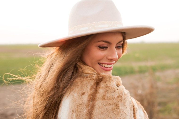 high school senior girl with long brown hair wearing a western hat wrapped in a brown blanket smiles over her shoulder standing in a field near OKC with the sunset behind her
