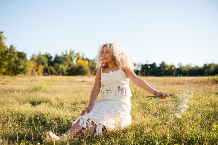 image of a high school senior girl with blonde curly hair wearing a white western boho fringe dress holding out a bouquet of baby's breath and looking away sitting in the middle of a field at Martin Nature Park in Oklahoma City