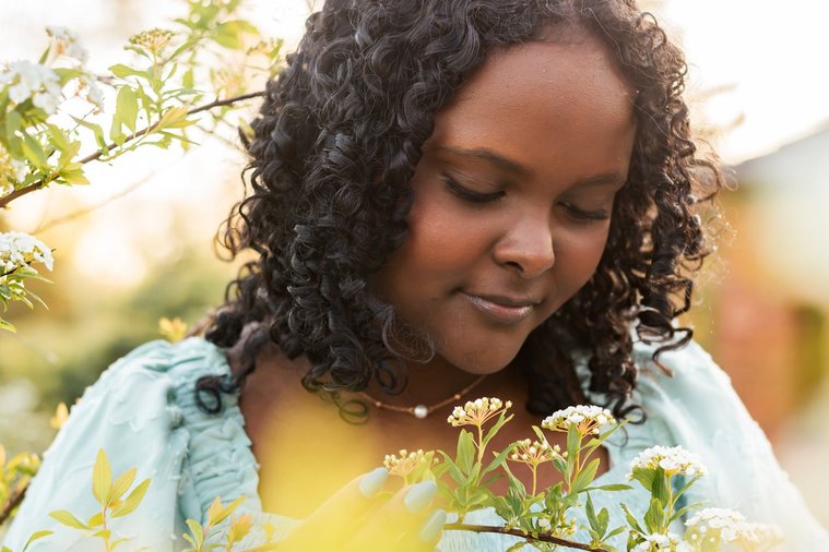 close up portrait of a black senior girl looking down at a flowering bush with green and flowers out of focus around her