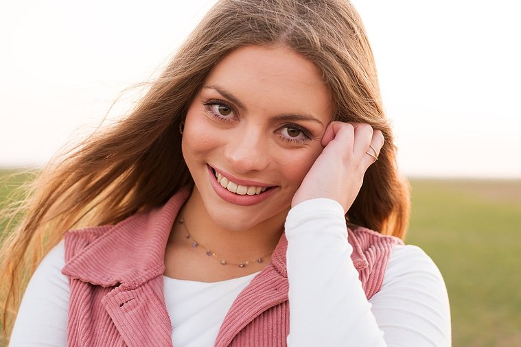 portrait of a beautiful senior girl with long hair wearing a pink vest smiling with one hand in her hair standing in front of a winter wheat field in the country near Oklahoma City