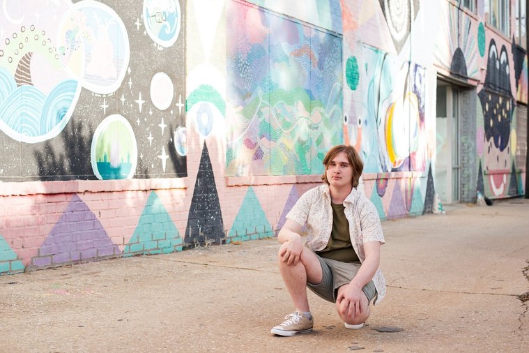 senior boy wearing a button down shirt and blue shorts squats in an alley with a serious face in front of a colorful wall mural in midtown Oklahoma City
