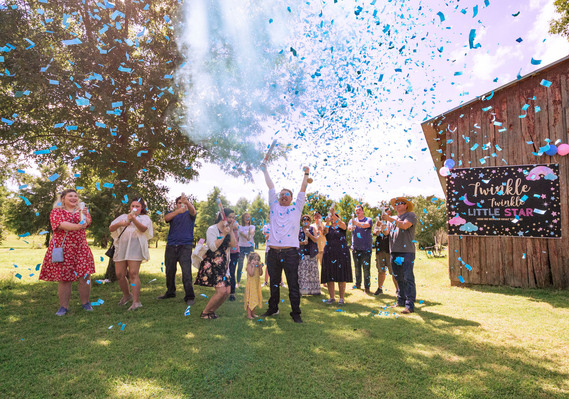 A large family group at a gender reveal with blue confetti and blue smoke cannons blowing around them.
