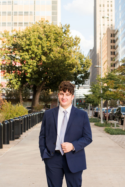 senior boy in a blue suit poses on a sidewalk in downtown OKC smiling with buildings and street behind him