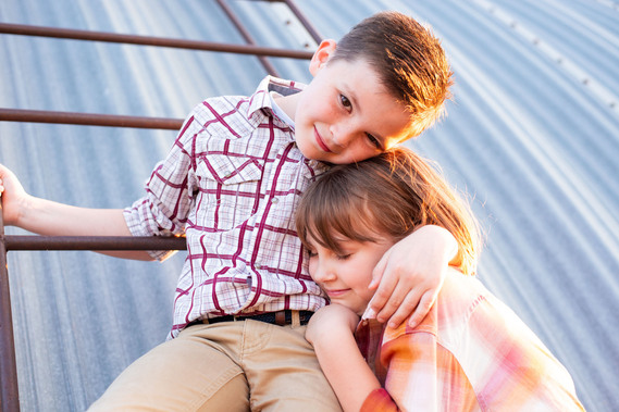 A brother and sister hug in front of a metal barn on a ladder at their back to school photo session on the farm.