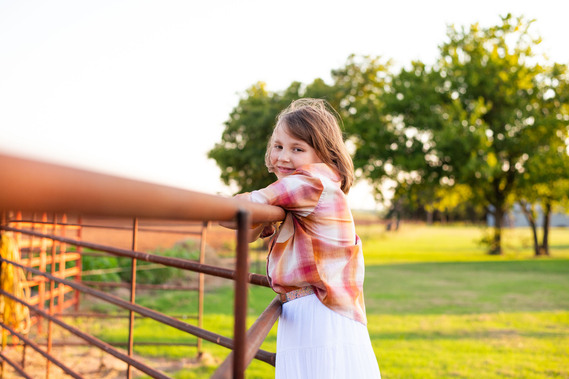 A little girl smiles as she stands on a metal fence at her back to school photo session in the country in Oklahoma