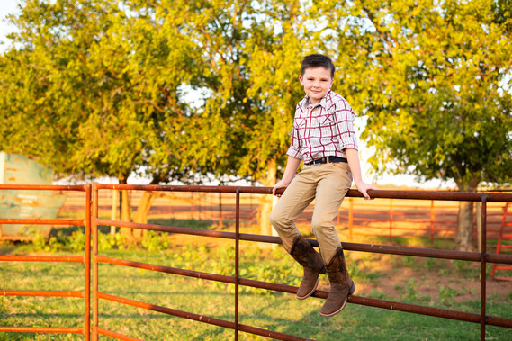 A little boy sits on a metal cattle fence in his cowboy boots in his back to school photo session in the country in Oklahoma
