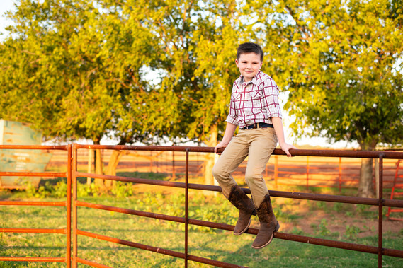 A little boy sits on a metal cattle fence in his cowboy boots in his back to school photo session in the country in Oklahoma