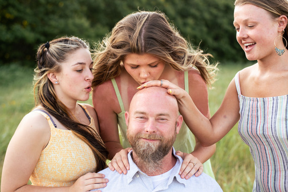 A photo of a father and his three teenage daughters who are messing with his bald head in front of a grassy field in rural Oklahoma at sunset