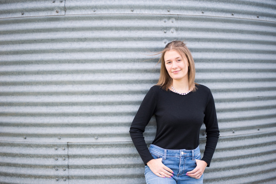 Portrait of a teenage girl in a black t-shirt and jeans standing in front of a grain bin with her thumbs in her pockets with a soft smile.