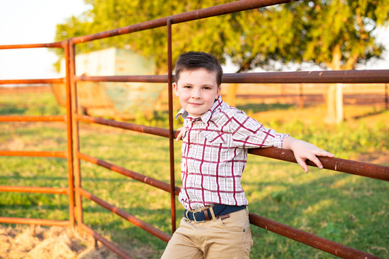 A little boy leans against a metal cattle fence and smiles in his back to school photo session in the country in Oklahoma