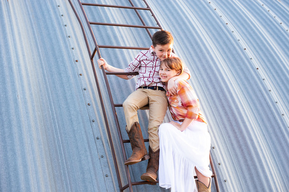 A brother and sister hug in front of a metal barn on a ladder at their back to school photo session on the farm.