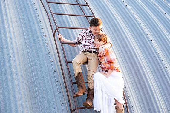A brother and sister hug in front of a metal barn on a ladder at their back to school photo session on the farm.