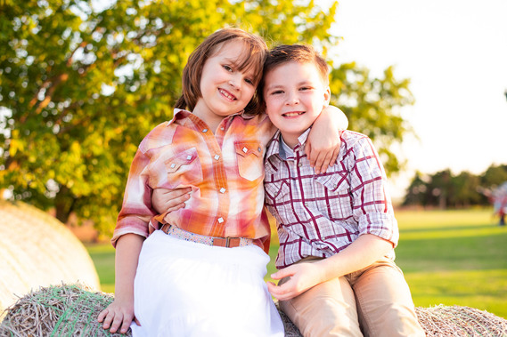 A brother and sister with their arms around each other are sitting outside in the country on a hay bale at their back to school photo session on the farm.