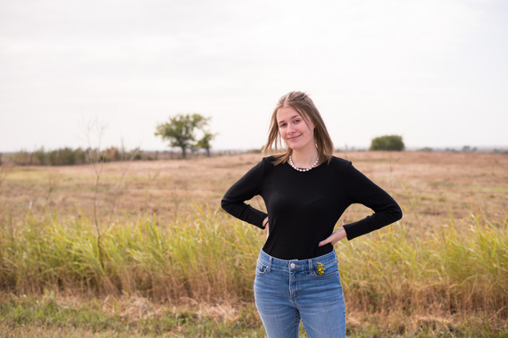 portrait of a teenage girl in a black shirt and jeans standing in front of a field with her hands on her hips