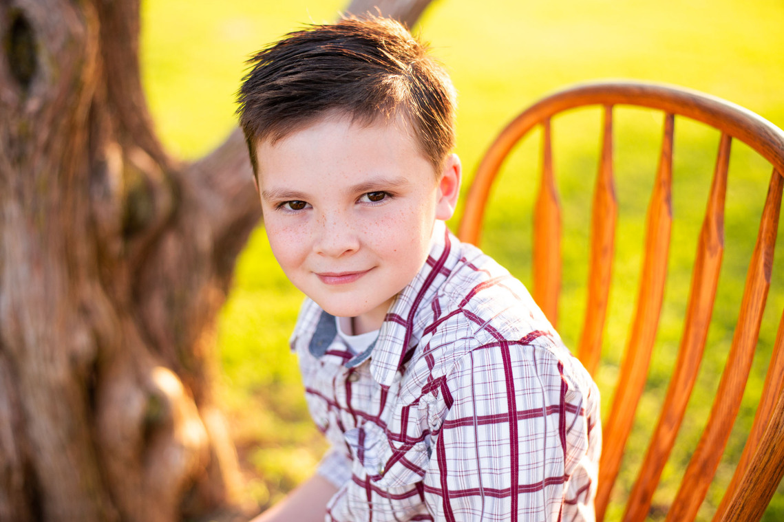 A little boy smiles while sitting in a chair in front of a tree at his back to school photo session in the country in Oklahoma