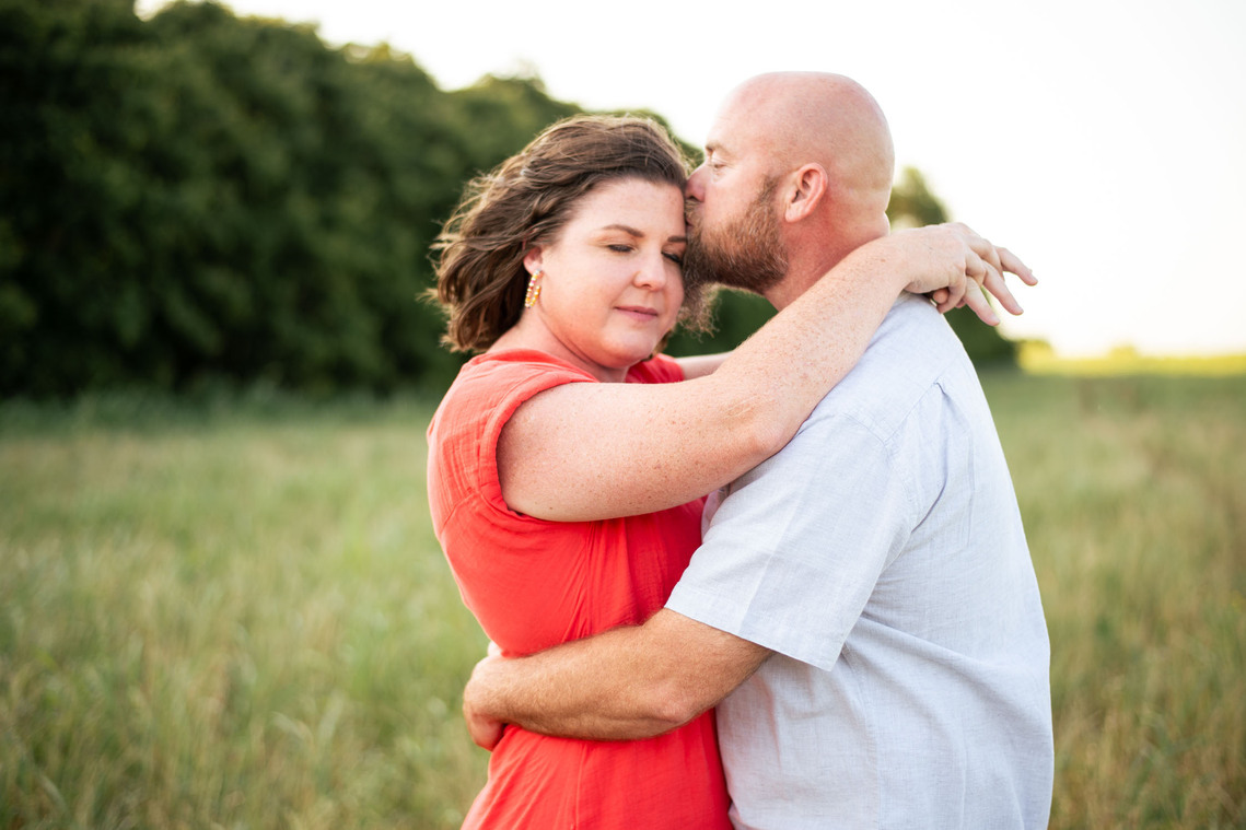 A husband kisses his wife's forehead in a photography pose in front of a flower meadow at sunset in Oklahoma
