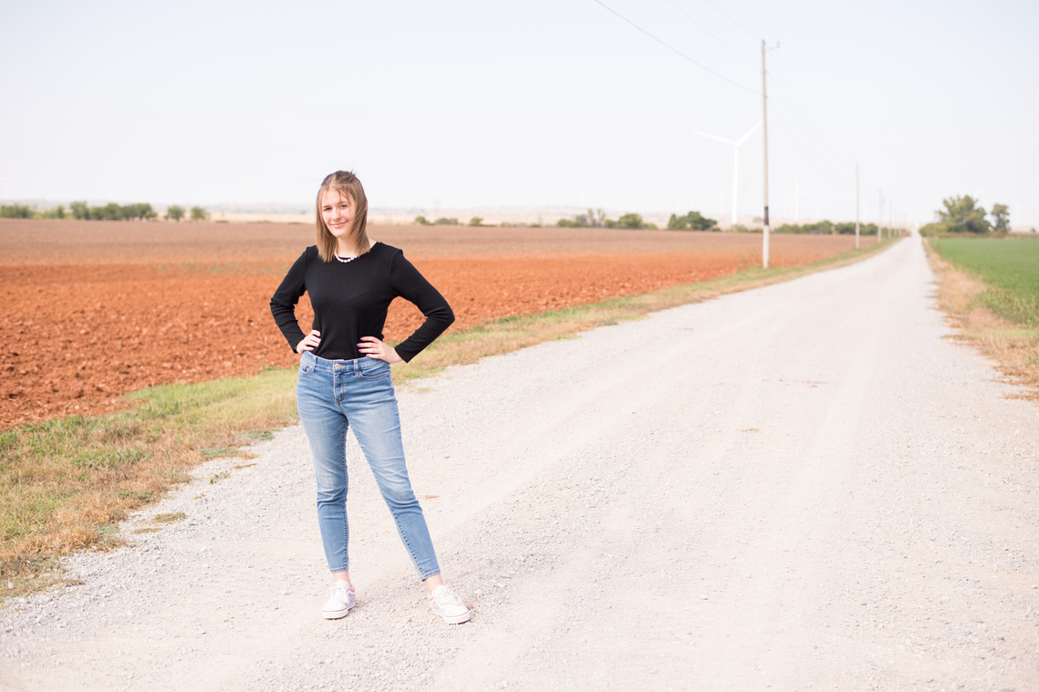 Portrait of a teenage girl in a black t-shirt and jeans standing in the middle of a dirt road with her hands on her waist.