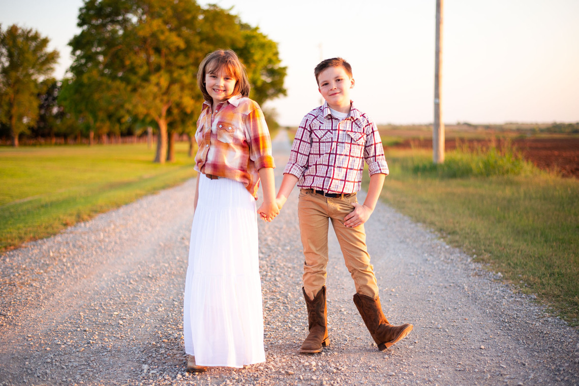 A brother and sister in country clothes hold hands and smile while standing in the middle of a gravel road at their back to school photo session on the farm.